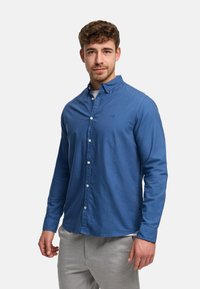 Nuori mies, jolla on lyhyet kiharat hiukset, hänellä on sininen nappipaita valkoisen aluspaidan päällä ja vaaleanharmaat housut, seisoo yksiväristä taustaa vasten.