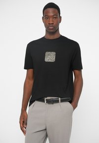 T-shirt preta de manga curta com um embellishment metálico quadrado no peito, combinada com calças texturizadas cinzentas e um cinto preto.