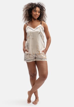 Woman standing barefoot with hands in pockets wearing beige patterned camisole and matching shorts against plain background.