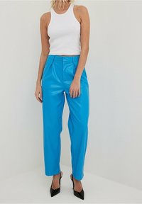 Blaue Kunstlederhose mit hohem Bund, gefaltetem Vorderteil, weiten Beinen und glattem, glänzendem Material; kombiniert mit einem weißen Top und schwarzen Absätzen.