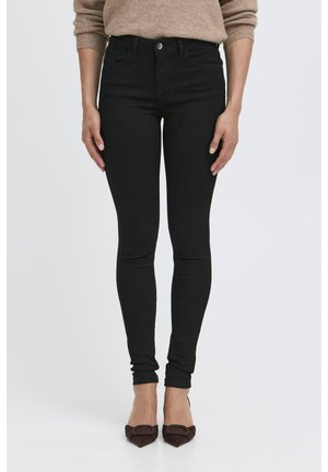 Jean skinny noir à texture lisse, coupe taille haute, avec une fermeture frontale à boutons, accompagné de chaussures marron à talons.