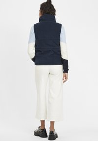 Marinblå kordflodsväst med hög krage, ihop med en flerfärgad randig tröja, vita vida byxor och svarta plattformsskor.