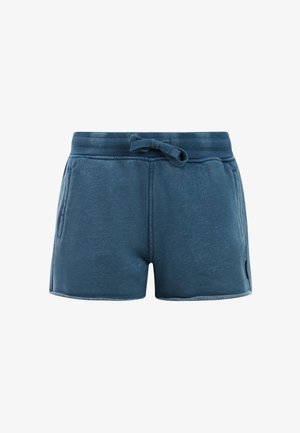 Meisje draagt een roze shirt, marineblauwe korte broek, roze sandalen, grote zonnebril en een roze haarband, staat met één hand in de zak.