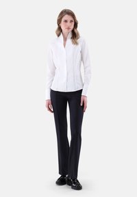 Weißes Hemd mit hohem Kragen und figurbetontem Design, kombiniert mit schwarzen geraden Hosen und schwarzen Schuhen. Einfacher, strukturierten Look.