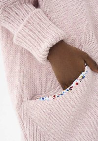 Jasnoróżowy sweter z dzianiny z żeberkowymi mankietami, z kieszenią wykończoną kolorowym kwiecistym wzorem. Miękka tekstura i luźny fason.