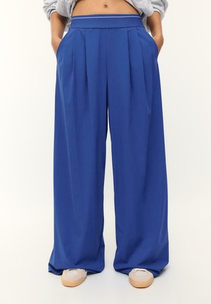 Person iført højtaljede, vide blå bukser med folder, hænderne i lommerne, kombineret med beige sneakers og kort grå top.