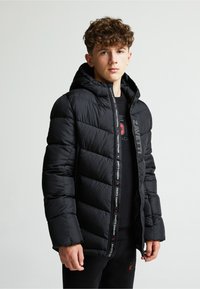 Melnā pūkainā jaka ar kapuci, ar quilted dizainu, logotipa akcentiem uz rāvējslēdzēja un matētu apdari. Valkāta virs melnas krekliņa.