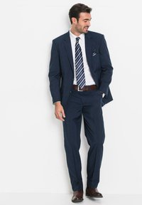 Abito blu navy con giacca su misura e pantaloni con gamba dritta, abbinati a una camicia bianca, una cravatta a righe e scarpe in pelle marrone.
