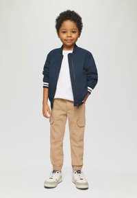 Marine bomberjack met witte strepen bij de manchetten, witte t-shirt, beige cargobroek en witte sneakers met grijze accenten.