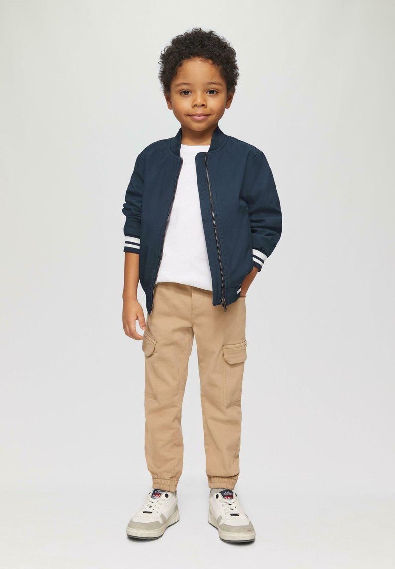 Marine bomberjack met witte strepen bij de manchetten, witte t-shirt, beige cargobroek en witte sneakers met grijze accenten.