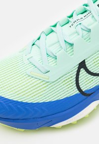 Chaussure de course vert clair et bleu avec une tige texturée en mesh, des lacets bien visibles et un logo noir audacieux sur le côté.