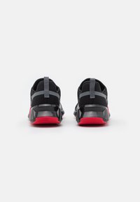 Een paar zwarte sportschoenen met rode zolen en grijze hielriemen, van achter bekeken op een witte achtergrond.