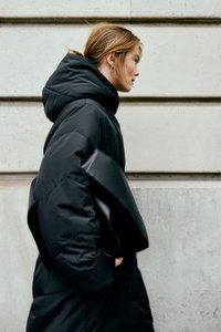 Una donna con i capelli corti indossa un grande piumino nero e orecchini a cerchio, mentre tiene in mano una grande borsa a tracolla nera contro un muro di pietra.
