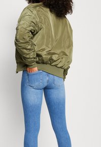Olivově zelená bombera s lesklým povrchem, žebrovanými manžetami a zipovou kapsou na levém rukávu, kombinovaná s modrými úzkými džíny.
