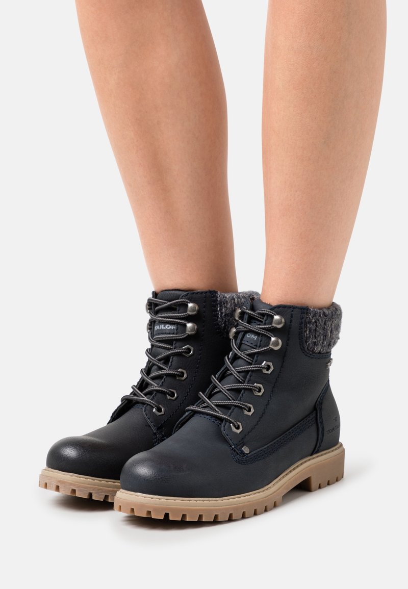 Bottes en cuir noir montantes jusqu'à la cheville avec poignets tricotés gris et semelles robustes beige portées sur des jambes nues.