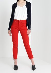 Rote Skinny-Jeans mit ausgefransten Säumen, kombiniert mit einem weißen Oberteil und einer marineblauen Strickjacke mit weißen Knöpfen, ergänzt durch schwarze spitze Schuhe.