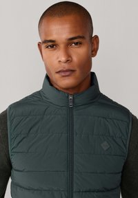 Donkergroene gewatteerde bodywarmer met een opstaande kraag en een ritssluiting aan de voorkant. Heeft een diamanten logo aan de linkerkant, met een horizontale doorgestikte textuur.