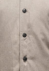 Μπεζ πουκάμισο με κουμπιά, featuring μια υφή σε σχέδιο ψαροκόκαλο και μαύρα κουμπιά. Η κοντινή άποψη τονίζει τις λεπτομέρειες του υφάσματος και των κουμπιών.