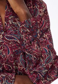 Arm gekruist, gekleed in een langwerpig shirt met een levendig rood, blauw en beige paisley-patroon en een knoopmanchet.