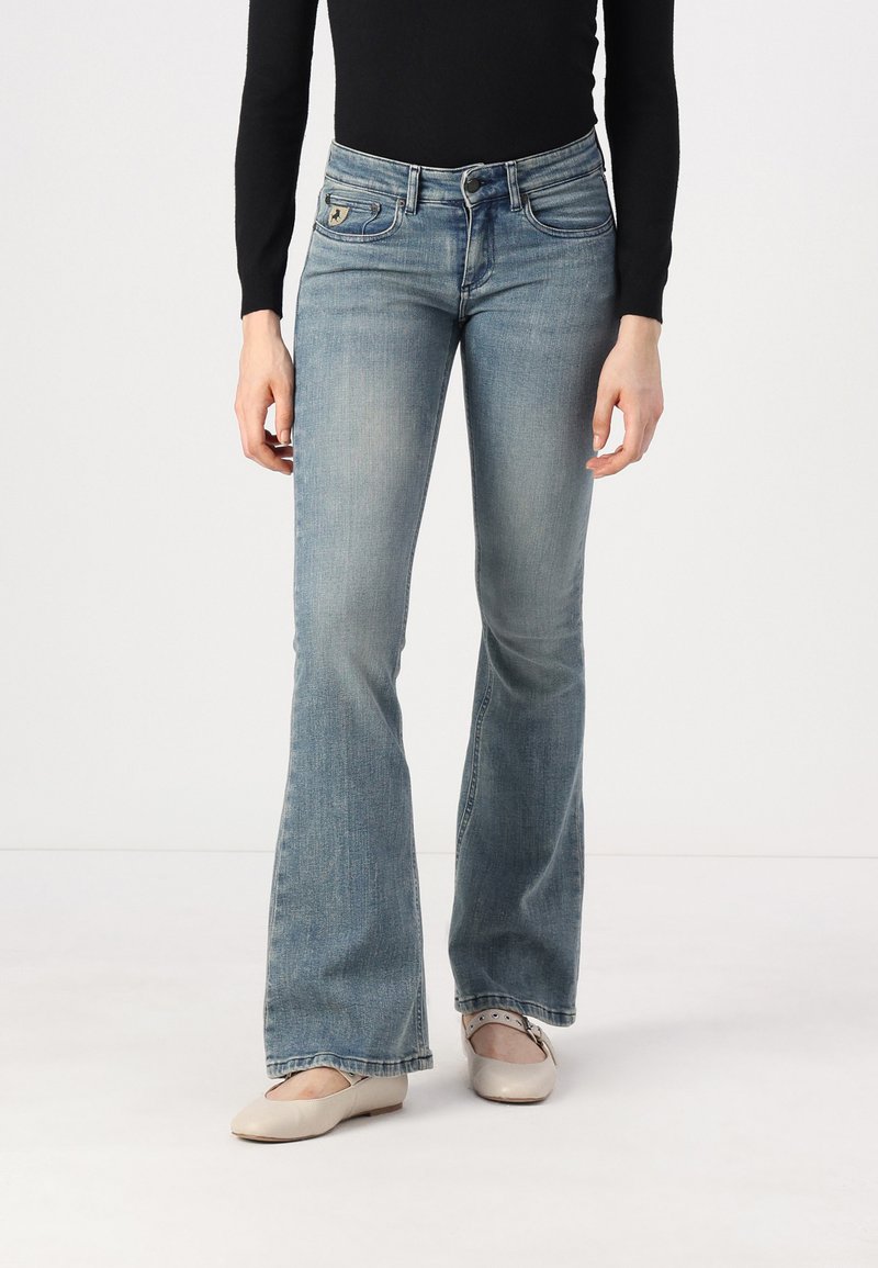 Lys blå bukser med vidde i denim, med frontlommer og et distinkt logo på livet. Glat tekstur med en let falmet overflade.