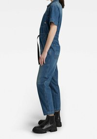 Combinaison en denim à manches courtes, avec des poches latérales et une ceinture noire. Présente une coupe décontractée et des jambes fuselées, assortie à des bottes noires à semelles épaisses.