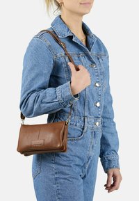 Donna in tuta di denim blu che tiene una borsa a tracolla in pelle marrone su sfondo bianco.