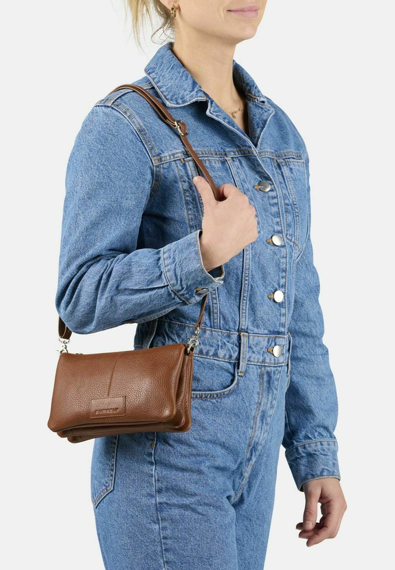 Donna in tuta di denim blu che tiene una borsa a tracolla in pelle marrone su sfondo bianco.