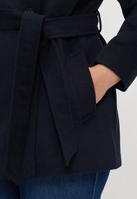 Navy-Wollmantel mit einem Gürtel, Seitentaschen und einer lockeren Passform. Der Stoff wirkt weich mit einer glatten Textur. Sichtbare Nähte entlang der Kanten.