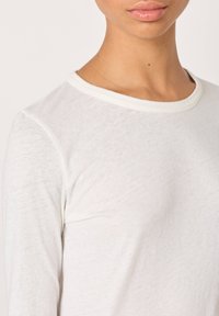 Valge pikkade varrukatega pluus ümmarguse kaelusega, valmistatud kergest kangast. Omaduseks on pehme tekstuur ja lihtne, puhas disain ilma mustriteta.