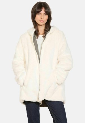 Cappotto in pelliccia sintetica bianca con zip frontale e cappuccio; presenta una texture morbida, vestibilità oversize e tasche laterali. Indossato sopra una maglia leggera e jeans blue.