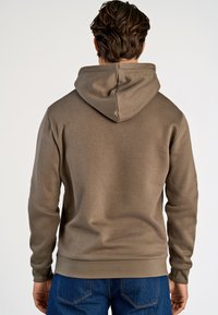 Sudadera marrón con una textura suave, que presenta una gran capucha y puños acanalados. Combinada con unos jeans azules, mostrando un ajuste casual.