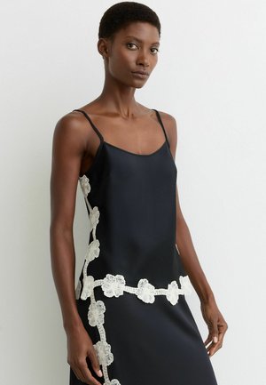 Kobieta ubrana w czarną, bezrękawnikową sukienkę z białą koronką w kwiatowy wzór wzdłuż boków i dołu, stojąca na tle jednolitej białej ściany.