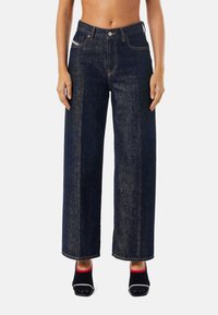 Džíny s širokými nohavicemi z tmavého denimu s vysokým pasem, kontrastním prošíváním a kapsami bez zapínání. Textura působí pevně a odolně.