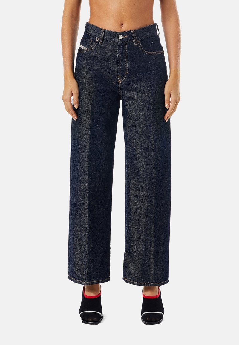 Džíny s širokými nohavicemi z tmavého denimu s vysokým pasem, kontrastním prošíváním a kapsami bez zapínání. Textura působí pevně a odolně.