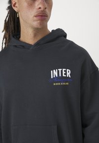 Sweatshirt escura com capuz, feita de uma mistura de algodão. Apresenta "INTER Nerazzurri" impresso em branco e azul, com um bolso frontal e um ajuste descontraído.