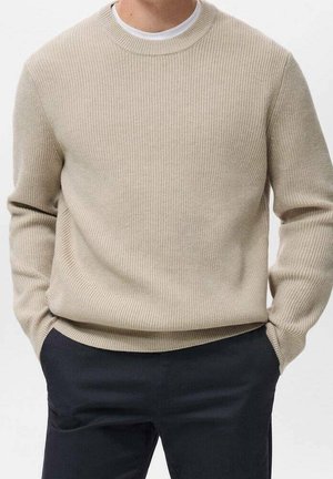 Mand iført beige ribstrikket sweater over hvid skjorte med hænderne i lommerne på mørke bukser mod ensfarvet baggrund.