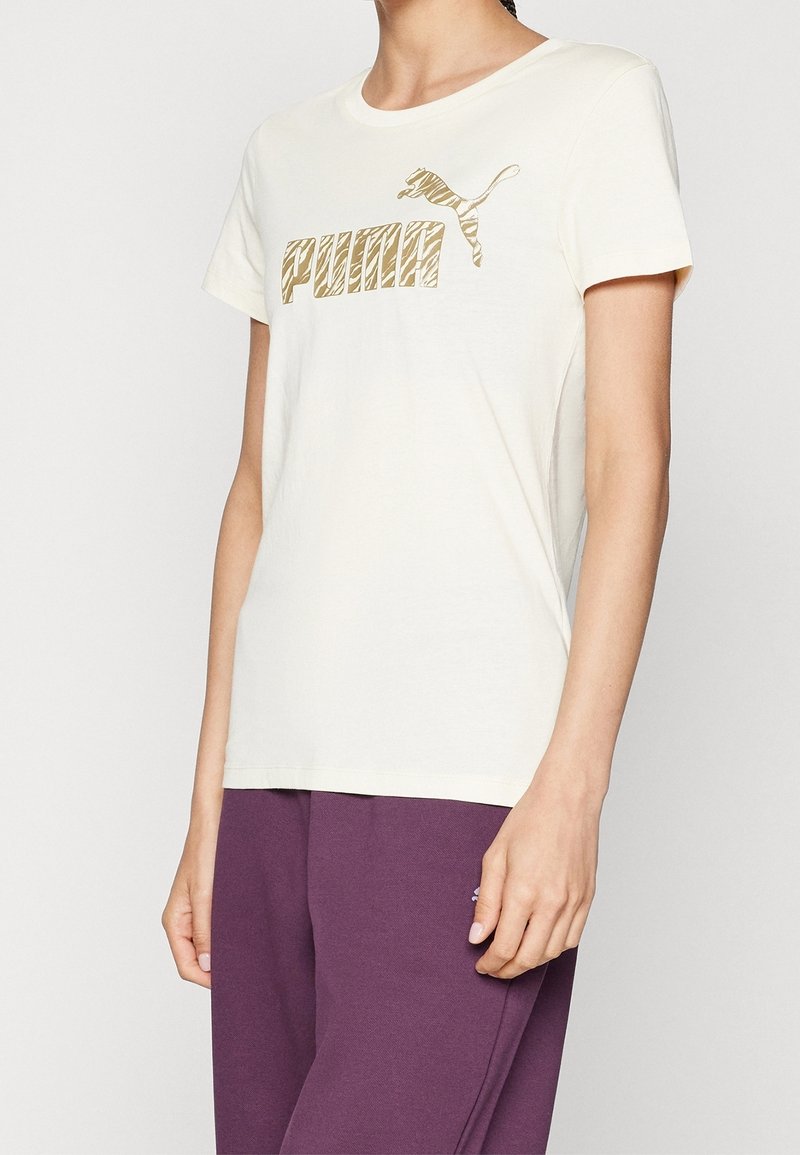 Kvinna i en krämfärgad Puma-t-shirt med guld zebrarandig logga och lila byxor, visad från axlarna till höfterna mot en enkel bakgrund.