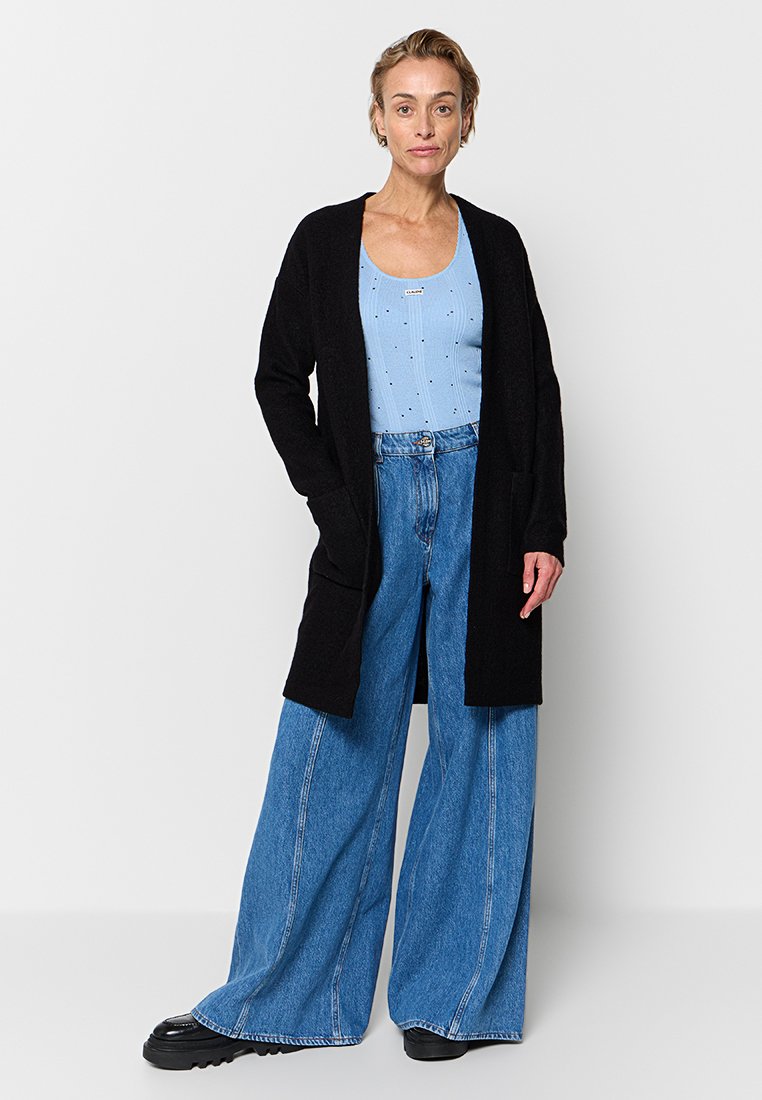 Schwarzer langer Cardigan mit offener Front, getragen über einem hellblauen Oberteil und weit geschnittenen blauen Jeans, mit einer weichen Textur und lässigem Fit.