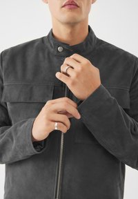 Giacca in pelle grigia con colletto, zip frontale e due tasche sul petto. Un uomo regola la manica con un anello d'argento su una mano.