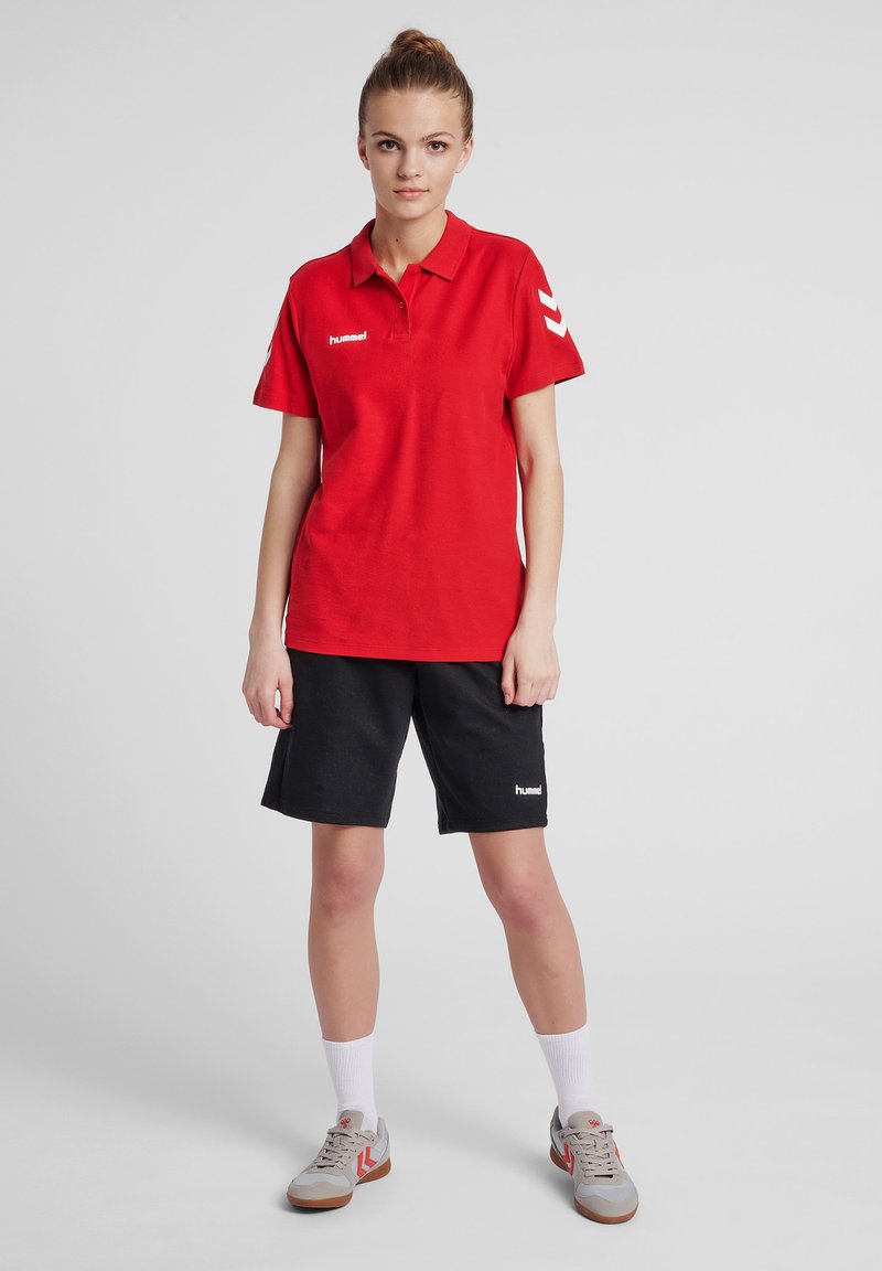 Rotes Poloshirt mit kurzen Ärmeln und weißen Akzenten; kombiniert mit schwarzen Shorts und grauen Sneakers. Aus Baumwolle, lässiges sportliches Design.