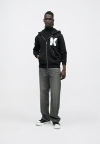 Sweatshirt preta com fecho de correr e gráfico branco, jeans cinzentos e ténis pretos. O conjunto apresenta um corte descontraído e texturas suaves.