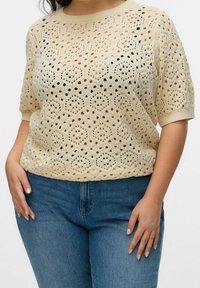 Donna che indossa un top corto in cotone a crochet beige abbinato a jeans in denim blu, mostrata dalla testa ai fianchi su uno sfondo bianco uniforme.