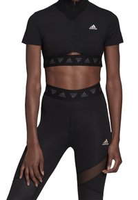 Černý sportovní crop top s vysokým límcem a výřezovými detaily, doplněný o černé legíny s síťovanou částí a jemnými akcenty značky.