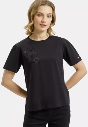 Blondynka w czarnej koszulce z krótkim rękawem z delikatnym kwiatowym haftem na lewej piersi, stoi z rękami za plecami.