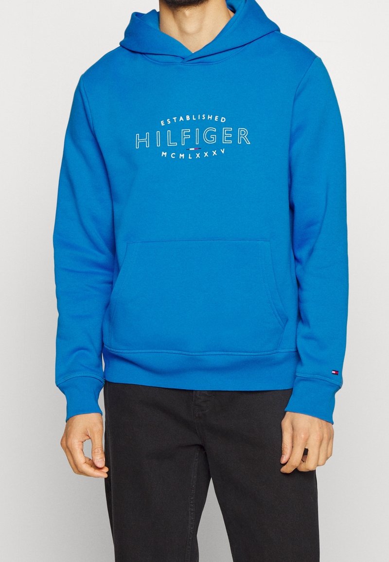 Homme portant un sweat à capuche bleu vif avec poche avant et texte blanc « Hilfiger Established MCMLXXXV » sur la poitrine, associé à un pantalon noir.