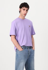 Light purple cotton T-shirt with short sleeves, crew neck, and a small logo patch featuring red, white, and blue accents. Worn with blue jeans.
