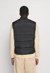 Svart pufferväst med horisontella quiltade sektioner, hög krage och lätt design, bärs över en beige sweatshirt.