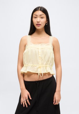 Jonge vrouw met lang zwart haar draagt een bleekgeel kanten mouwloos crop top en zwarte losse broek tegen een egale achtergrond.