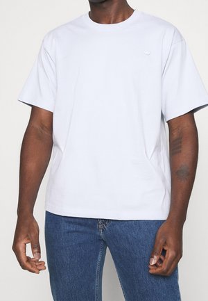 Jasnoszara bawełniana koszulka z okrągłym dekoltem, krótkimi rękawami i subtelnym logo na piersi. Noszona z niebieskimi dżinsami.