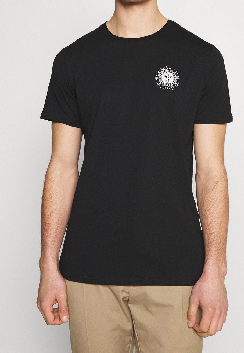 Homme portant un t-shirt noir avec un petit motif blanc représentant le soleil et la lune sur la poitrine, associé à un pantalon beige.